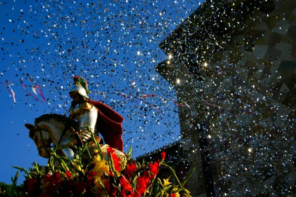 Cavaleiro em parada festiva com cavalo adornado, armadura dourada e manto vermelho.