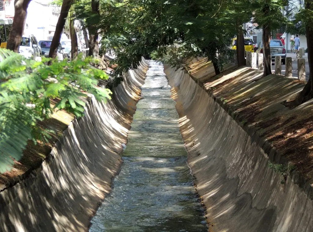 Canal de drenagem urbano com árvores e pilares de concreto.