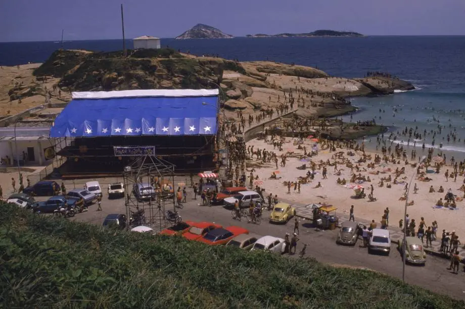 Praia lotada com palco e ilha ao fundo.