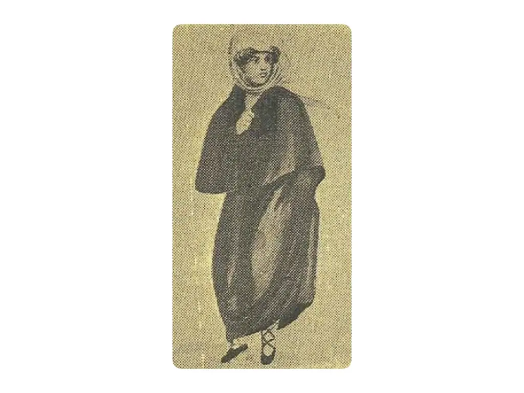 A figura feminina veste um josezinho, capa sem mangas feita em tecido vermelho. Na cabeça, ela usa lenço de cambraia engomada.