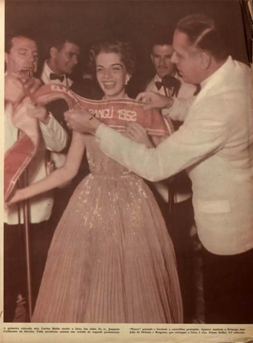 A senhorita Corina Baldo, de 21 anos, jovem funcionária da Embaixada Americana, foi a primeira colocada no concurso e recebeu a faixa de Miss Elegante Bangu de 1952 das mãos de Joaquim Guilherme da Silveira. Corina, que mais tarde se tornaria mãe do ator Felipe Camargo, usa vestido de organdi na cor mauve plissado e bordado com canutilhos. Ecktachrome de Nicolau Drei e Yllen Kerr, Manchete, 07/02/1953